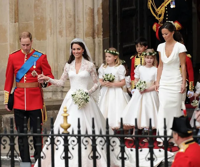 Royal wedding procession with bride in lace gown, groom in red uniform, and bridesmaids in white dresses.
