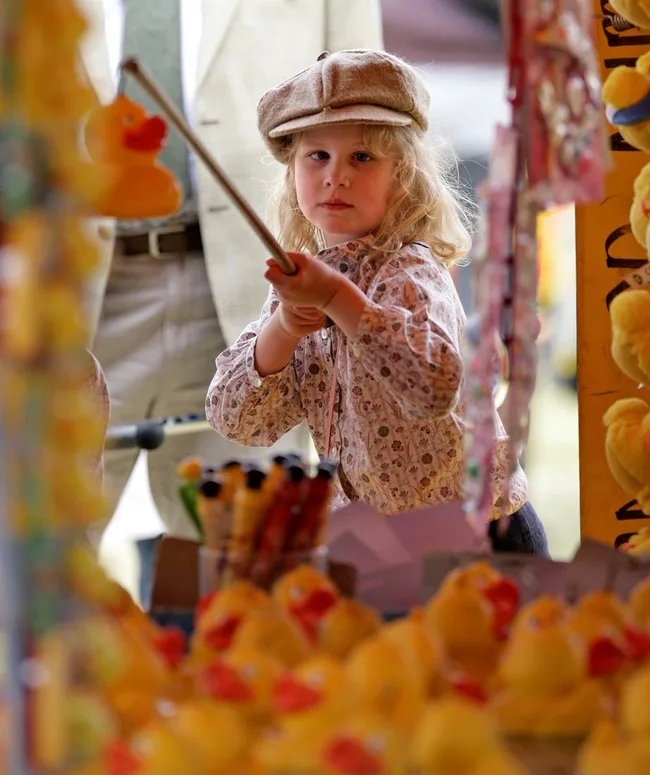 Child wearing cap at carnival game, holding pole to catch rubber ducks, focusing intently.