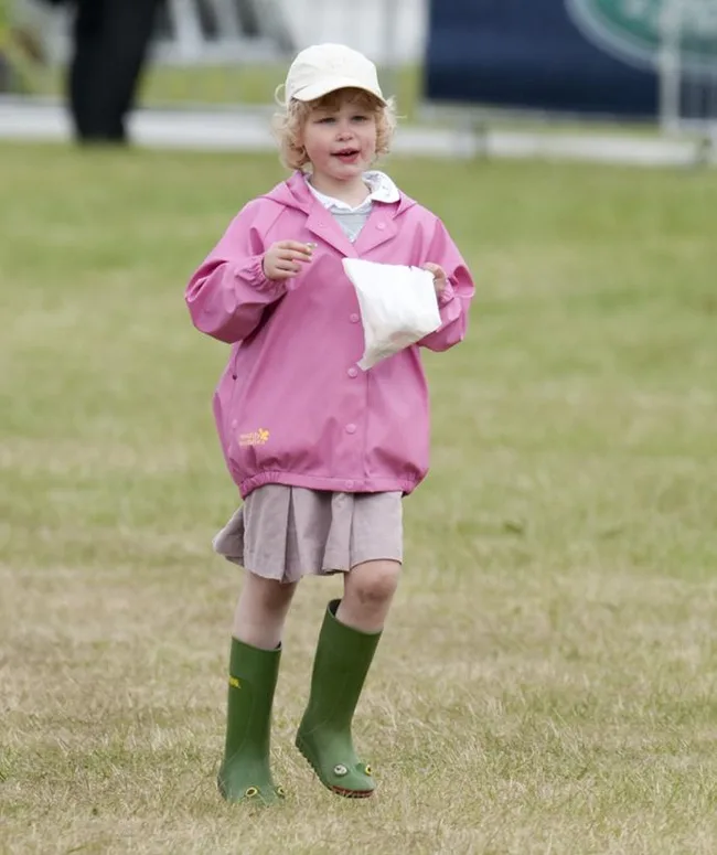 A young child in a pink coat and green boots holding a bag, walking on grass.