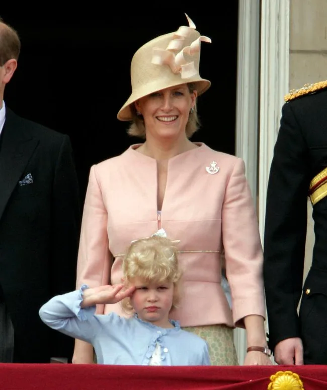 A woman in a beige hat stands with a young girl saluting; both are dressed formally at an event.