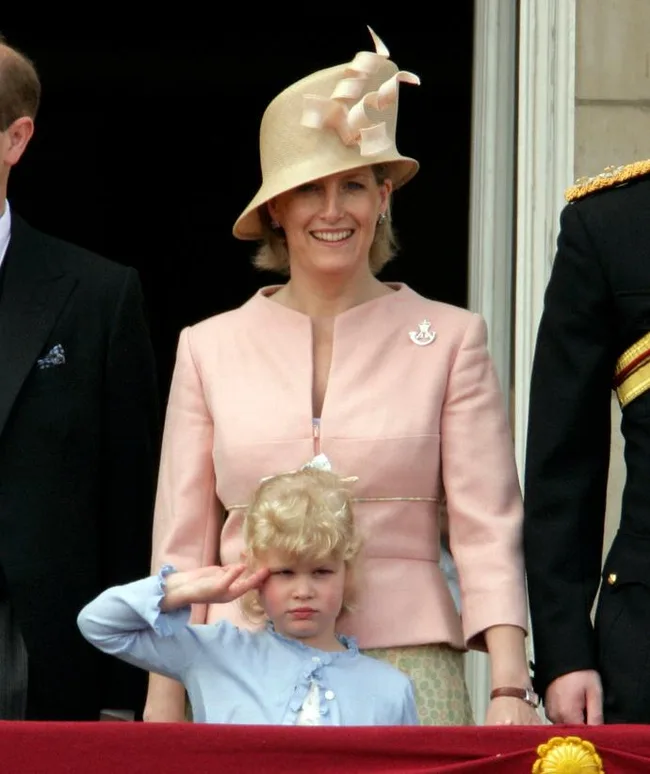 A woman in a beige hat stands with a young girl saluting; both are dressed formally at an event.