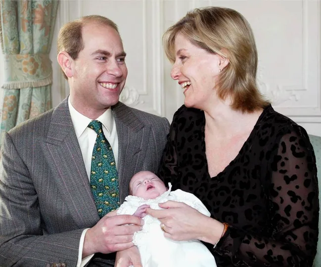 A smiling couple with a newborn baby wrapped in white clothing, sitting in a cozy room.
