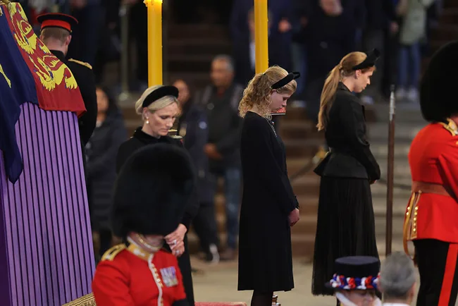Three women in black attire bow their heads in respect beside a draped coffin guarded by soldiers in ceremonial uniform.
