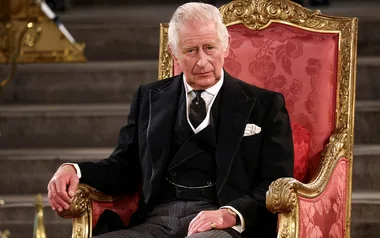 Man in a formal suit sitting on a gold-trimmed red throne-like chair, looking ahead with a neutral expression.