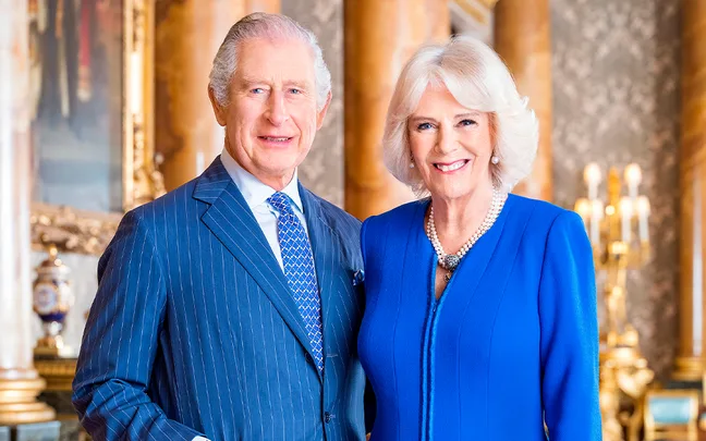 Two individuals smiling, wearing formal attire, standing in a richly decorated room with chandeliers and artwork in the background.