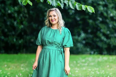 A woman in a green dress stands outdoors on grass, smiling with trees in the background.