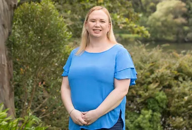 A woman in a blue shirt stands smiling outdoors in a green, leafy setting.