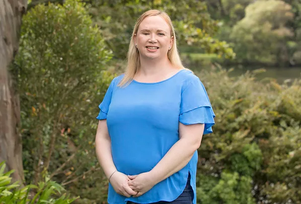 A woman in a blue shirt stands smiling outdoors in a green, leafy setting.
