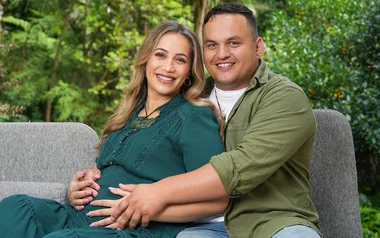 Couple sitting on a couch outdoors, with woman smiling and holding her baby bump, embraced by the man.