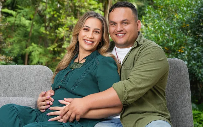 Couple sitting on a couch outdoors, with woman smiling and holding her baby bump, embraced by the man.