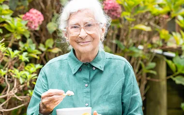 Elderly woman in a green shirt smiling, holding a bowl and spoon in a garden setting with blooming flowers in the background.