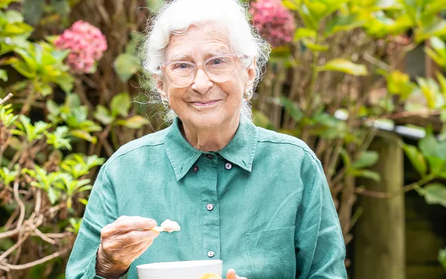 Elderly woman in a green shirt smiling, holding a bowl and spoon in a garden setting with blooming flowers in the background.