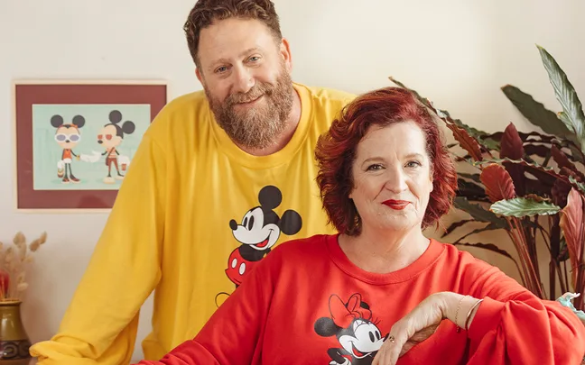 Two people in matching Mickey and Minnie Mouse shirts, smiling, with a framed artwork and plants in the background.