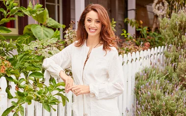 ```plaintext
Woman smiling in garden, standing by a white picket fence, wearing a light-colored blouse, amid green leafy plants.
```