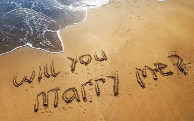 "Will you marry me?" written in sand with approaching ocean waves nearby.