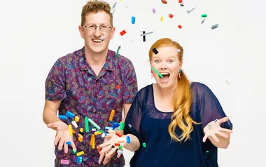 Two people joyfully throw colorful LEGO bricks into the air against a white background.