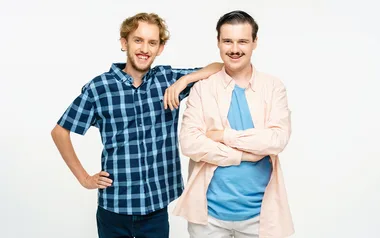 Two men smiling; one in a blue plaid shirt with hands on hips, the other in a pink shirt with folded arms.