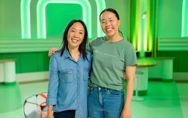 Two people smiling and posed together in a green-lit room, one wearing a denim shirt and the other a green T-shirt.