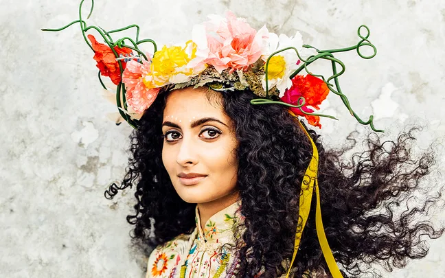 Woman with curly hair wears a colorful flower crown and patterned outfit, standing against a light textured background.