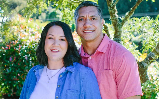 A couple smiling outdoors, under a tree with lush green foliage in the background.