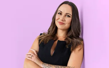 A woman with long hair and a tattoo smiles and crosses arms, wearing a black top against a light purple background.