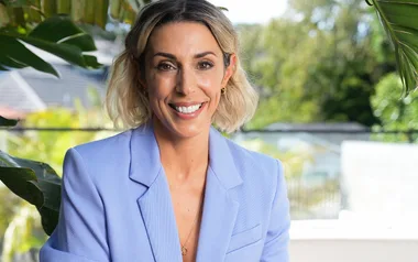 A smiling woman with short blond hair in a light blue blazer, outdoors with greenery in the background.