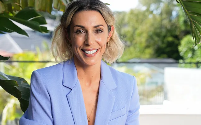 A smiling woman with short blond hair in a light blue blazer, outdoors with greenery in the background.