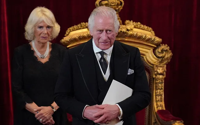A man in formal attire stands in front of a throne, with a woman in black dress by his side, both in a regal setting.