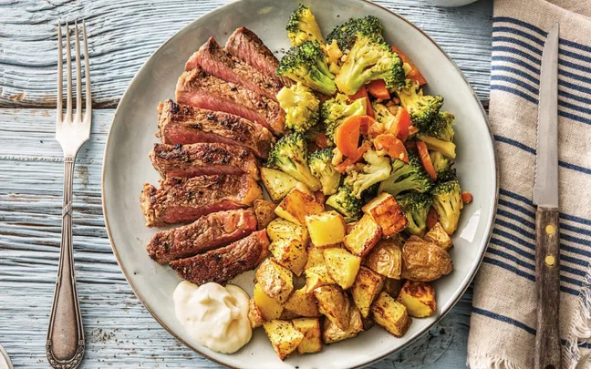 Sliced steak with sautéed broccoli and carrots, roasted potatoes, and a dollop of sauce on a plate.