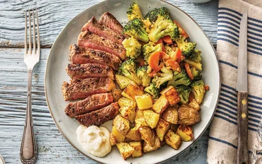 Sliced steak with sautéed broccoli and carrots, roasted potatoes, and a dollop of sauce on a plate.