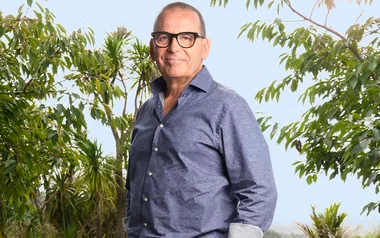 Man in glasses and blue shirt stands outdoors surrounded by green trees.