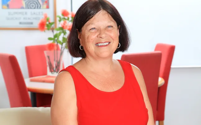 A woman in a red sleeveless top smiling indoors with flowers and chairs in the background.