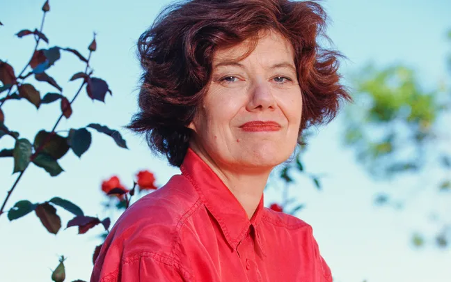 A person with short brown hair in a red shirt stands outdoors near roses, looking into the distance.