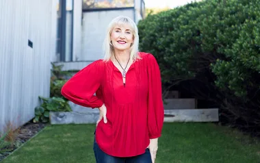 Blonde woman in a red blouse stands outdoors, smiling with hand on hip.