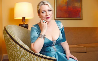 A person in a teal gown sits in an armchair, with a lamp and painting in the background.