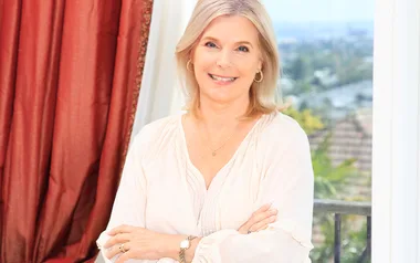 A smiling woman with blonde hair stands near a window with a scenic view in the background.