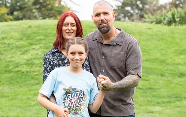 A family of three stands in a grassy field, smiling at the camera.