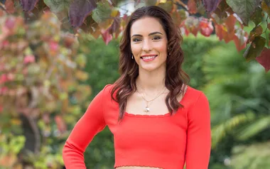 A woman in a bright orange top stands outdoors, surrounded by colorful autumn leaves.