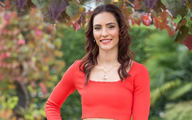 A woman in a bright orange top stands outdoors, surrounded by colorful autumn leaves.