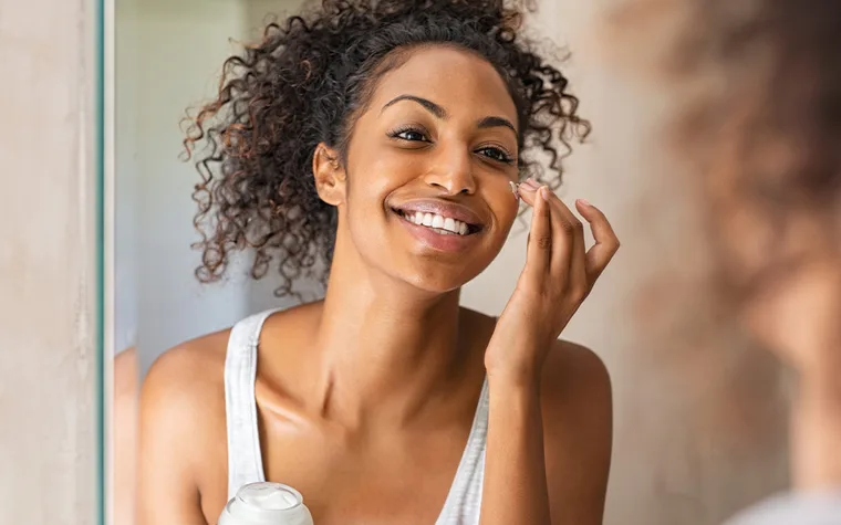 Smiling woman applying cream to face in front of a mirror.