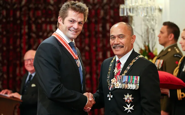 Two men in formal attire shaking hands at an award ceremony, both wearing medals and looking at the camera.