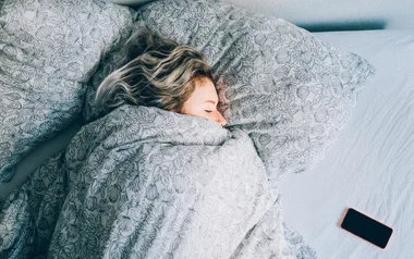 Person with blonde hair sleeping under a floral duvet, smartphone on the bed beside them.