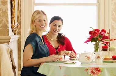 Two women smiling, seated at a floral-patterned table with a tea set, cupcakes, and a vase of red flowers.