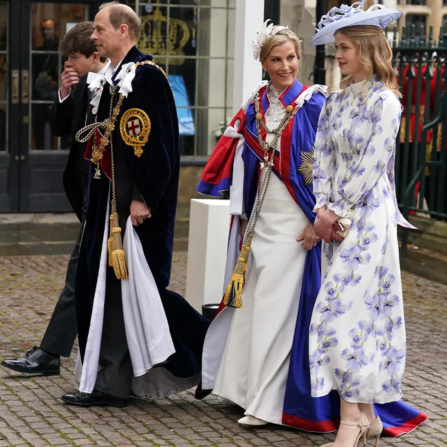 Four people walk together; two in ceremonial attire and two in formal wear, including a floral dress and hat.