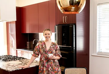 Woman in a floral dress stands in a modern kitchen with maroon cabinets and a marble countertop, holding a cup.