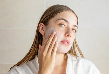 Woman applying facial cleanser, wearing a white robe, in a bathroom setting.