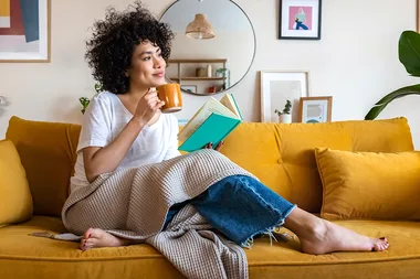 Person with curly hair reads a book and drinks coffee on a yellow couch, surrounded by home decor.