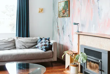 Cozy living room with a gray sofa, blue curtains, abstract wall art, fireplace, and a potted plant on wooden floor.