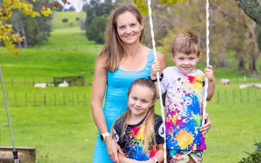 A woman and two children on a swing in a lush park, all smiling.