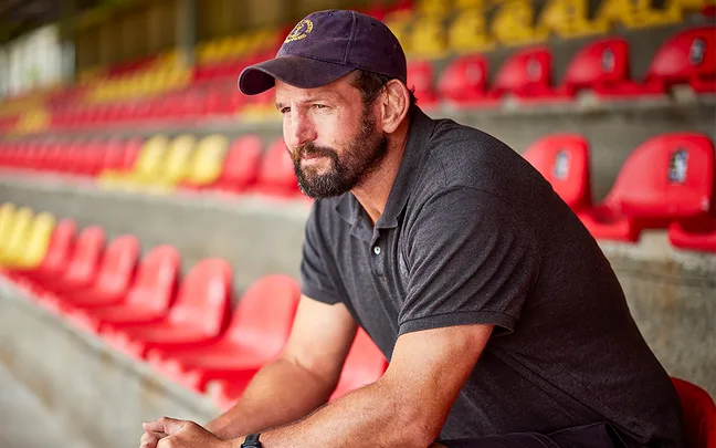 Carl Hayman in a black polo shirt and navy blue cap sits in a rugby stadium looking out to the field